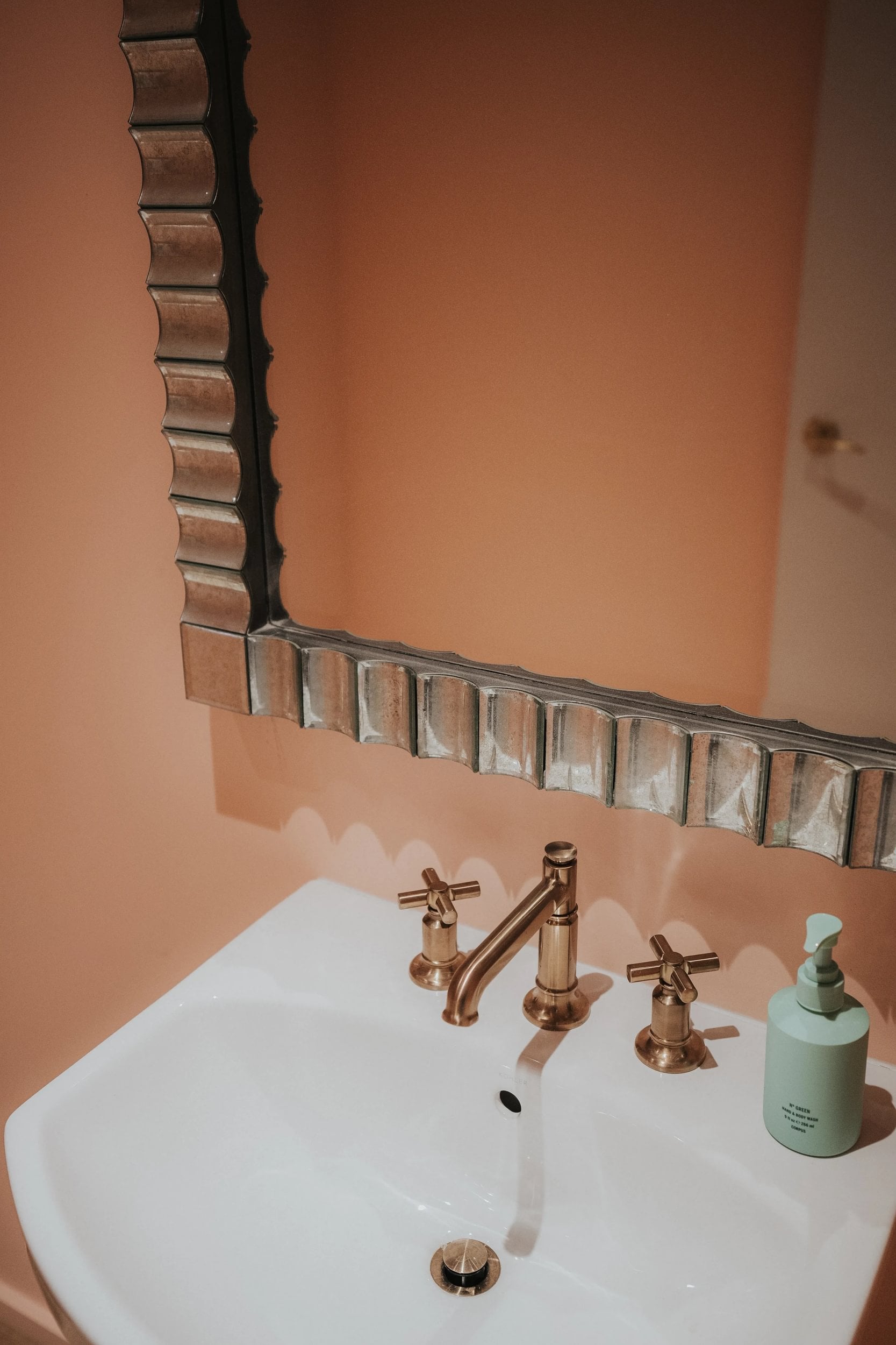 A close-up of a bathroom mirror and sink area with peach walls and gold fixtures.