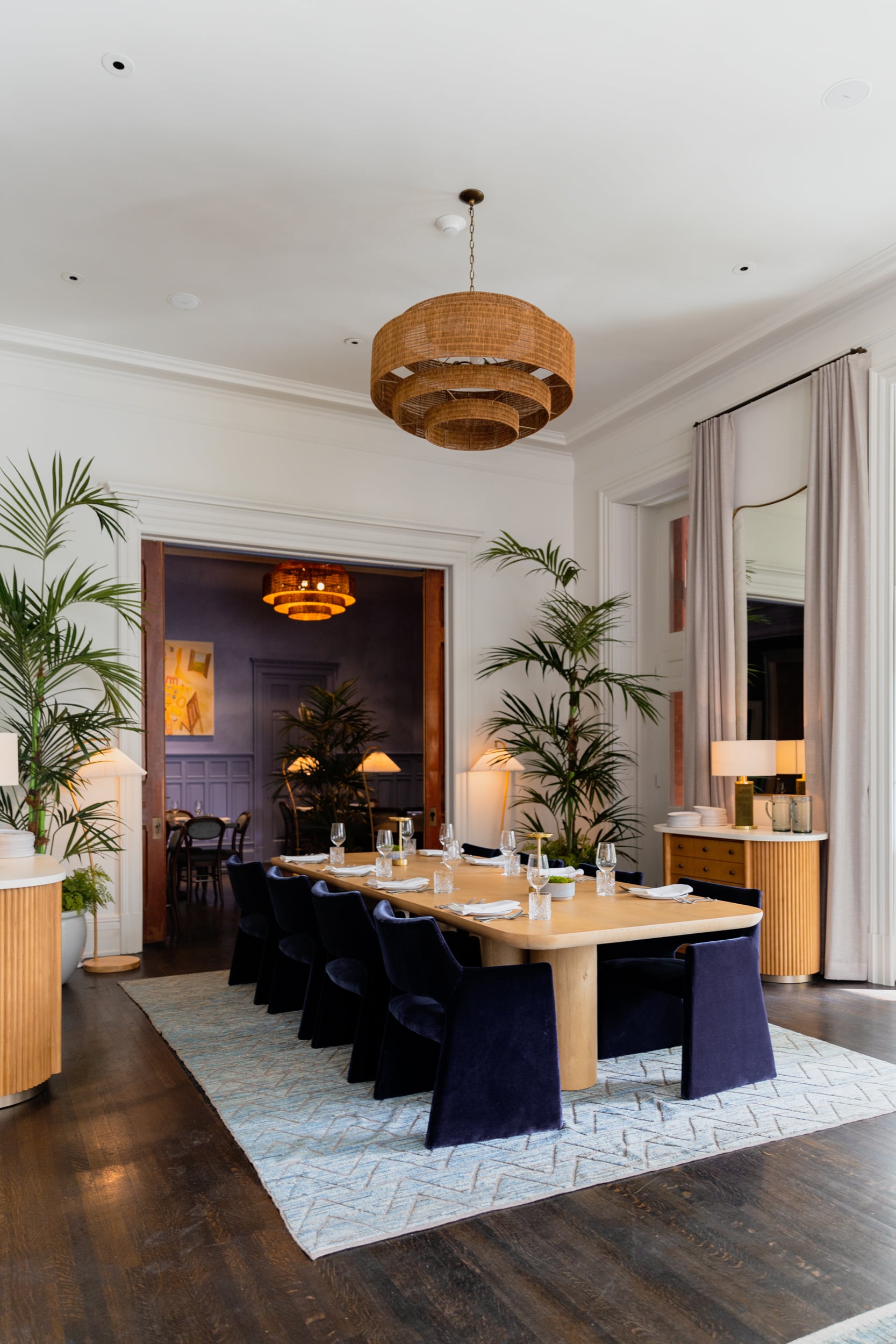 A dining room with a long table, chairs, plants, and lighting fixtures.