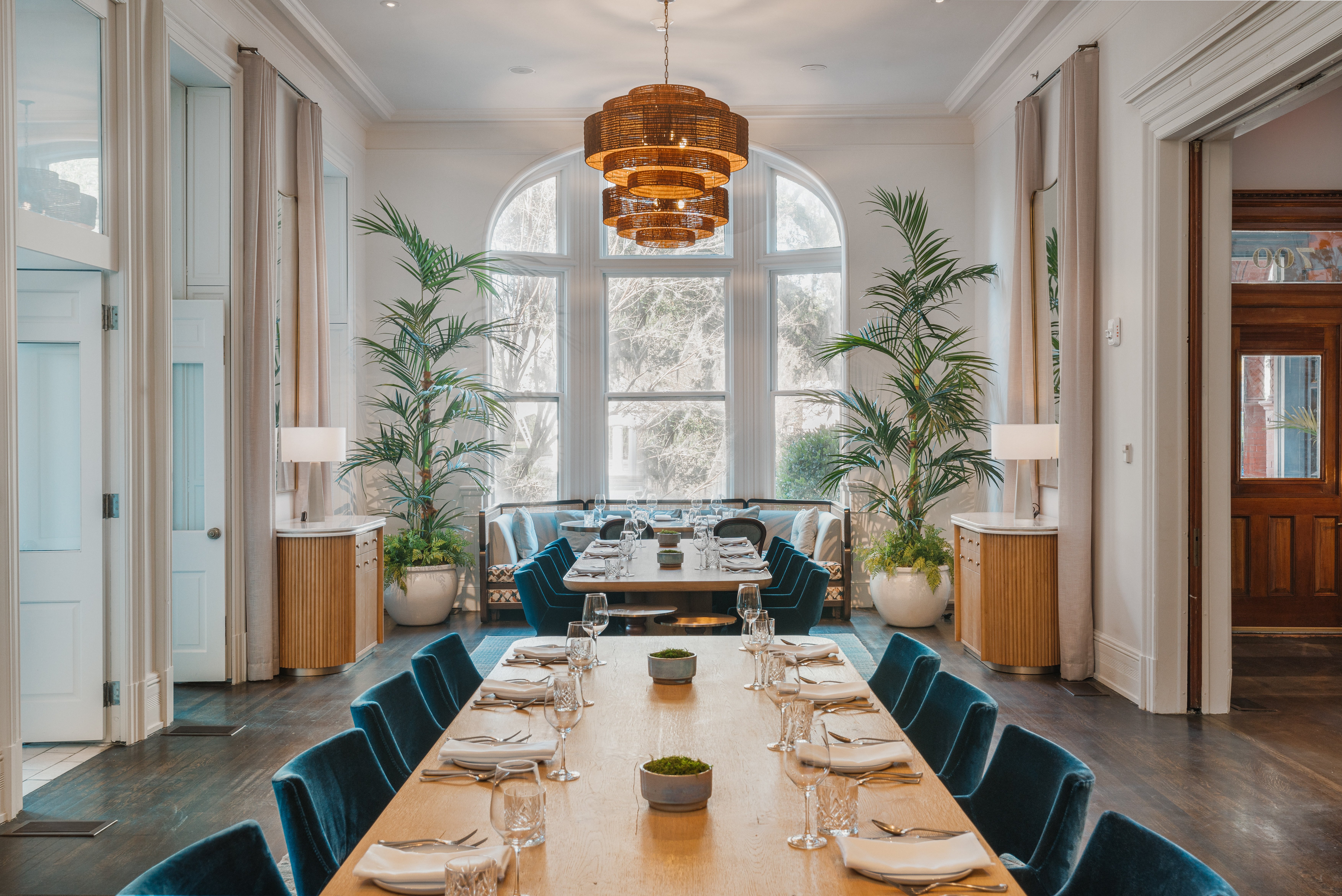 A dining room with a long table set for a meal, blue chairs, a large window, and plants.