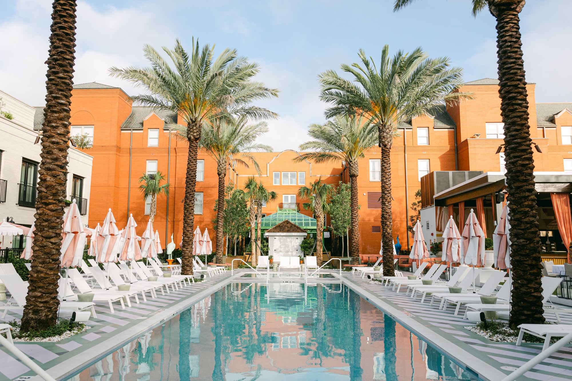 A pool area with white lounge chairs and palm trees in front of an orange building.