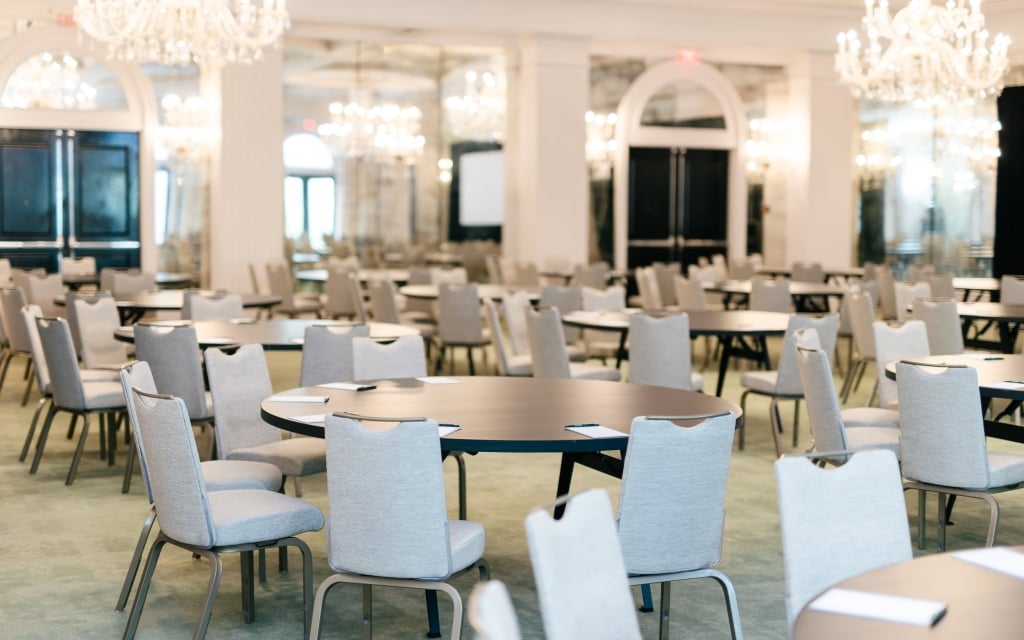 A large meeting room with round tables, white chairs, and chandeliers.