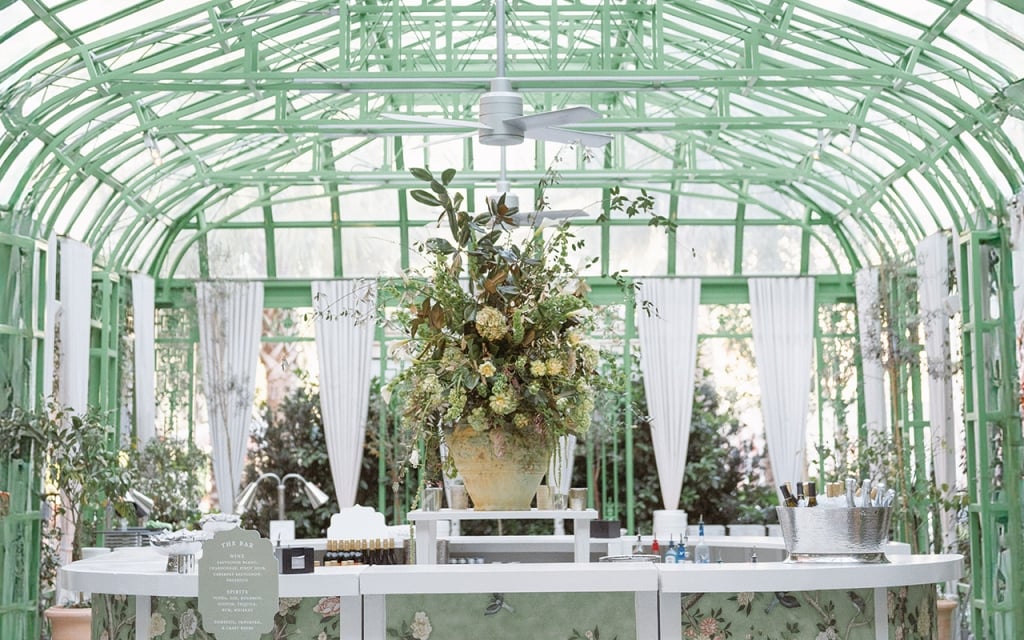 A large, bright room with a green metal frame, glass panels, and white curtains, featuring a large floral arrangement on a table.