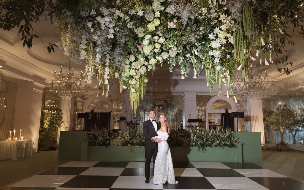 A couple in formal attire standing on a black and white checkered floor under a large floral arrangement in a luxurious lobby.