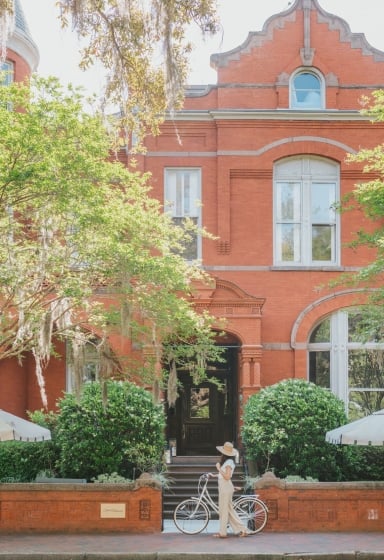 A red brick building with white trim and an arched doorway, with a woman walking with a bicycle in front of it.