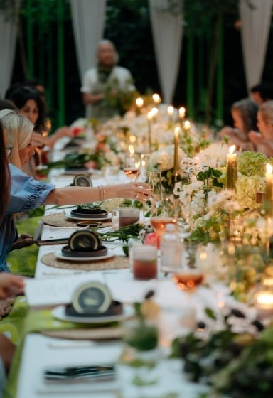 A long table set for dinner with people seated along both sides, under candlelight and surrounded by flowers.