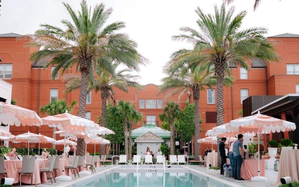 A poolside area with tables, chairs, and umbrellas, surrounded by palm trees and a large red brick building.