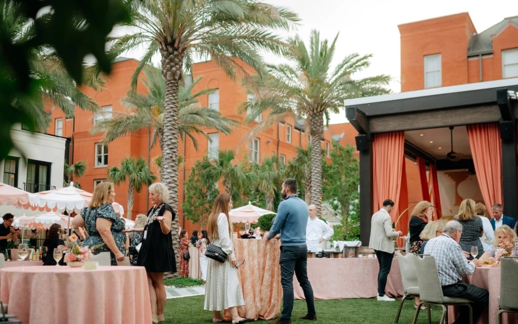 A group of people are gathered in an outdoor event space with palm trees, tables with pink tablecloths, and orange curtains.