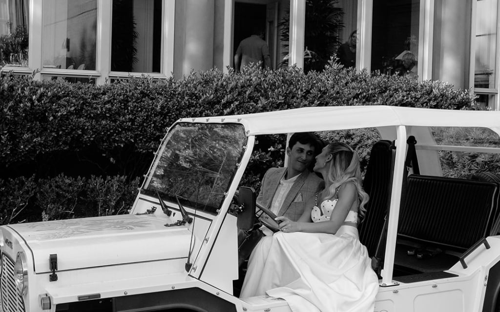 A black and white photo of a couple in a white jeep in front of a building.