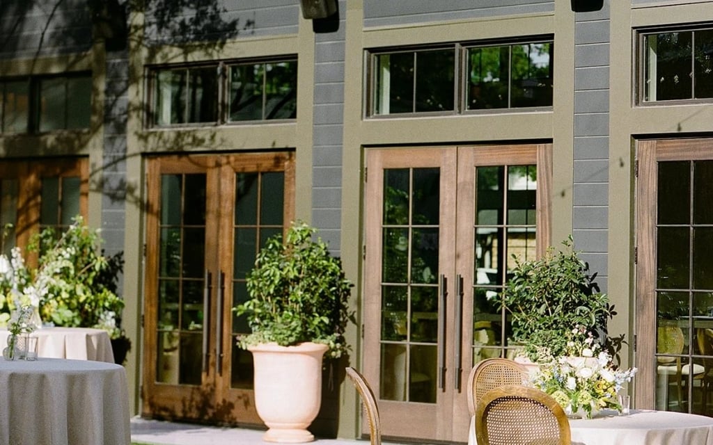 A serene outdoor event space with tables, chairs, and plants in front of a building with large doors and windows.