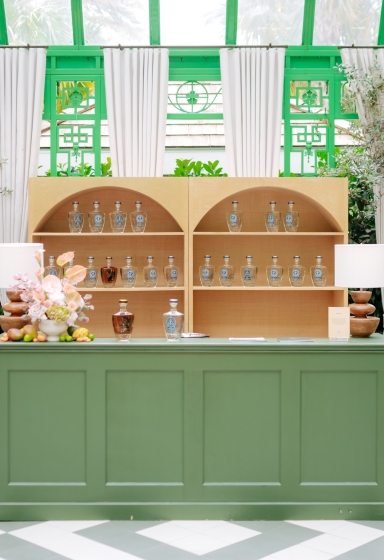Green bar station with arched bottle display shelving inside a lush botanical greenhouse with rattan lamps and café seating.