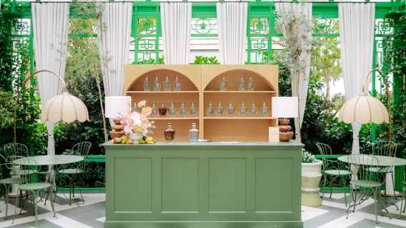 A green bar with tables and chairs, shelving with bottles, and lamps in a garden setting.