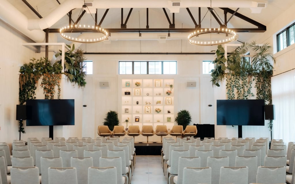 A well-lit meeting room with rows of chairs, a stage with seating, two large screens, and decorative plants.