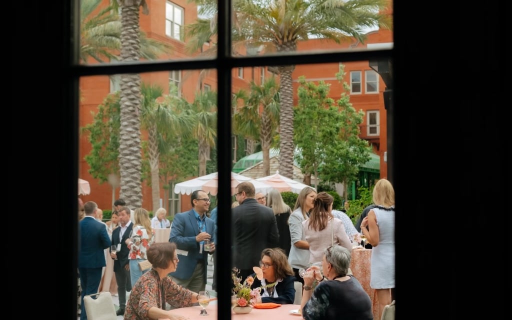 A group of people gathered at tables in an outdoor courtyard with palm trees and umbrellas.