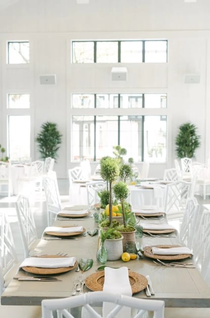 Carriage House event hall set for dining with rattan chargers, topiary centerpieces, and floor-to-ceiling windows, all in white