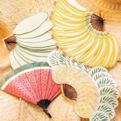 Four decorative folding fans shaped like tropical fruits — watermelon, banana, lemon, and pineapple — on a woven straw surface.