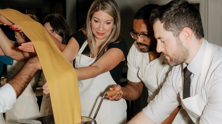 Guests all wearing white aprons help each other flatten homemade pasta via stainless steel pasta press